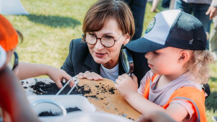 Zwei Kinder untersuchen gemeinsam mit Bildungsministerin Bettina Stark-Watzinger und einer Lupe ein Häufchen Erde auf einem Holztisch